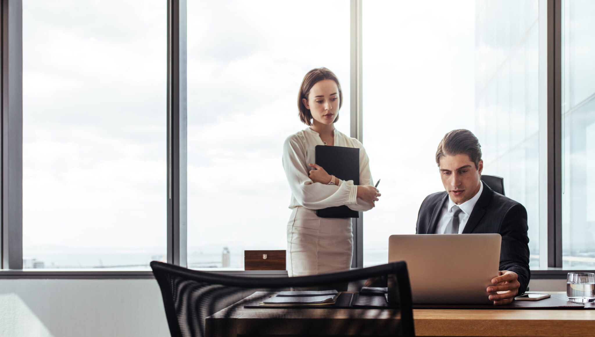 Two business people looking at laptop while working in the office. Businessman sitting at his desk with female coworker standing by.